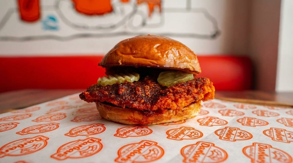 A crispy fried chicken sandwich with pickles on a branded wrapper, with a red booth in the background.