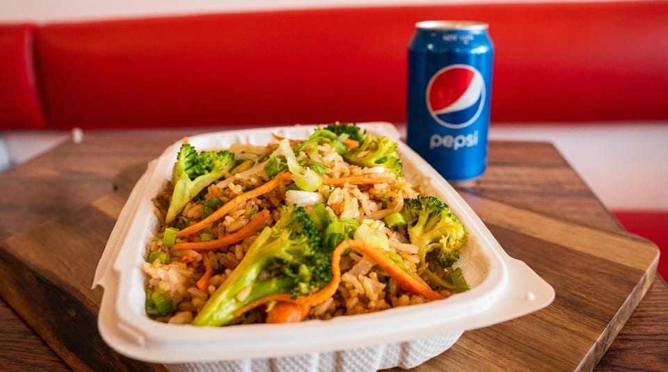 Fried rice with broccoli, carrots, and scallions in a takeout container with a Pepsi can.